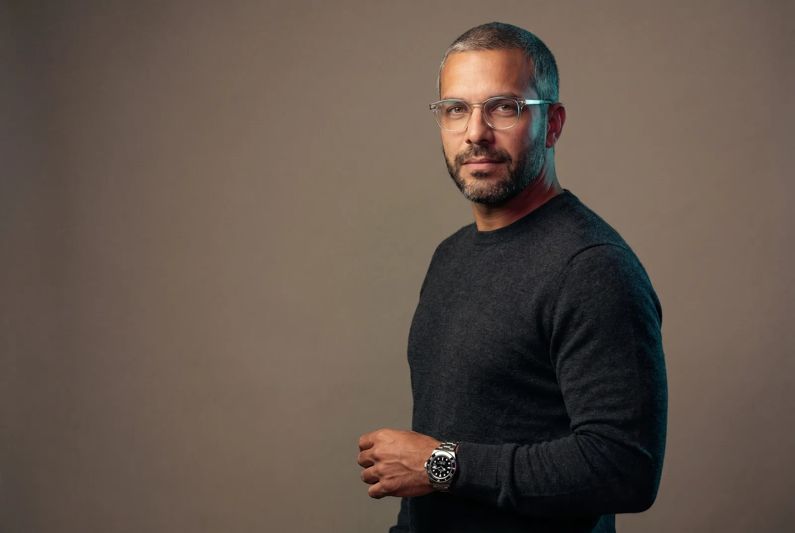 AI avatar generator concept shown as one custom AI avatar — a mid-40s spokesperson in clear-frame glasses and a steel dive watch — presented to camera as the single saved identity reused across stills, video, and ads, rendered by the Playcut Actor Engine