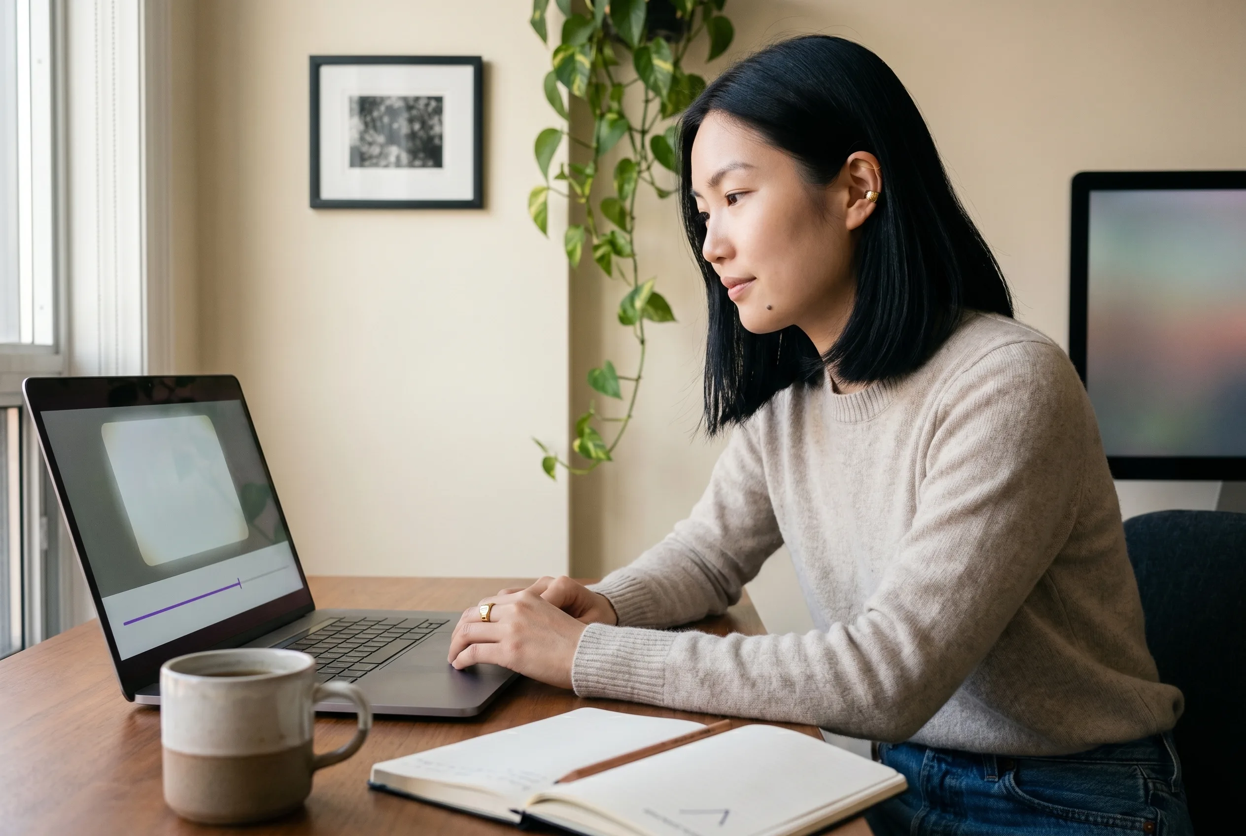 Mira Chen at her Toronto home-office workstation in the moment her first generated Playcut video plays back on her laptop, hands resting at the keyboard, a thin violet timeline bar glowing beneath the playback frame, soft north-facing window light catching her gold ear cuff and signet ring, Playcut Imagen 4 generation.