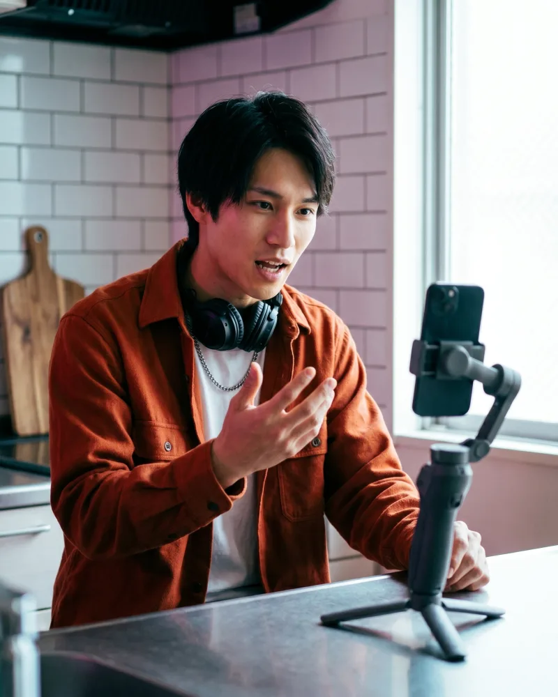The recurring AI actor mid-delivery of a new UGC hook to a phone on a small gimbal in a bright kitchen, wearing a different overshirt than earlier frames — a fresh hook fronted by the same saved face, rendered by the Playcut Actor Engine