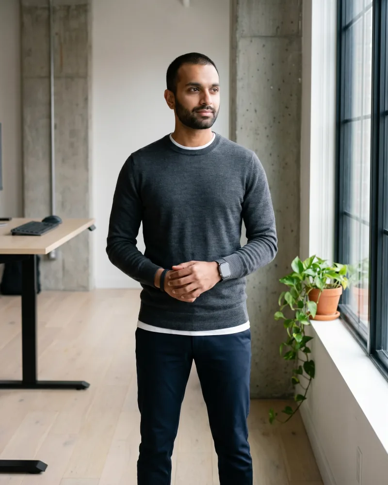 Editorial tech-founder portrait of Rohan, a South Asian man with a short trimmed beard and titanium smartwatch in a charcoal merino crewneck and slim navy trousers, standing near a black-framed industrial window in a modern minimalist office, Playcut Actor Engine generation