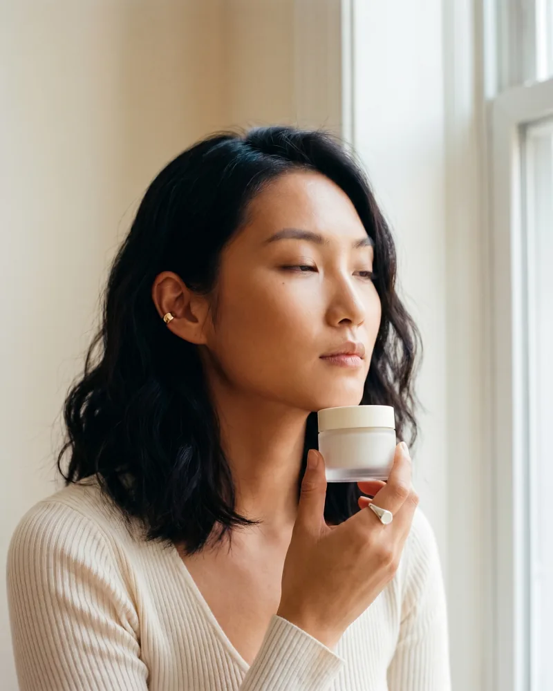 Editorial beauty still of the same custom AI actor Mira Chen holding an unbranded frosted-glass skincare jar at collarbone height in soft north-facing window light, ivory ribbed knit, Playcut Actor Engine generation