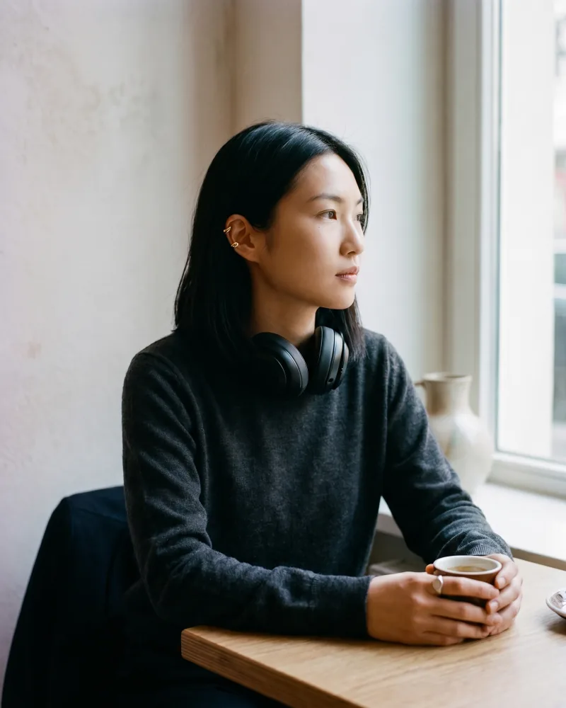 Editorial lifestyle still of the same custom AI actor Mira Chen at a café table with matte-black over-ear studio headphones resting around her neck, charcoal merino crewneck, espresso cup in hand, Playcut Actor Engine generation