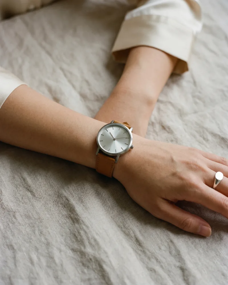 Editorial product-on-body macro still of the same custom AI actor Mira Chen's wrist wearing an unbranded brushed-steel watch with warm-tan leather strap on rumpled natural linen, signet ring visible, Playcut Actor Engine generation