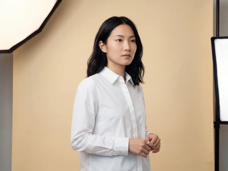 Clean studio softbox portrait of the same custom AI actor Mira Chen in a white cotton-poplin shirt against a seamless warm cream paper backdrop with even wraparound soft light, Playcut Studio Softbox filter