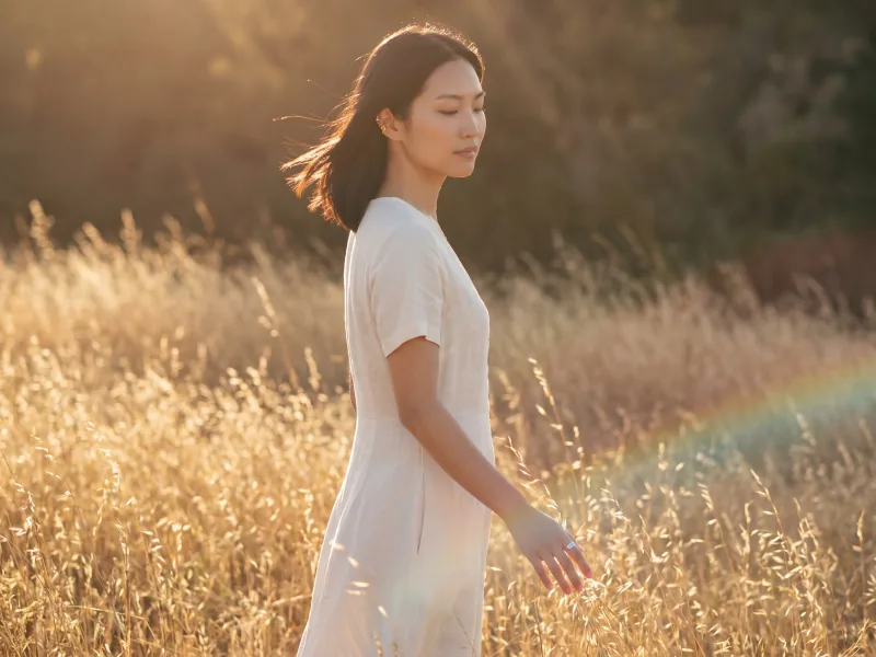 Golden-hour outdoor editorial photograph of the same custom AI actor Mira Chen in an ivory linen midi dress walking through a sunlit field of dry tall grass with warm sun backlight rim-lighting her hair, Playcut Golden Hour filter