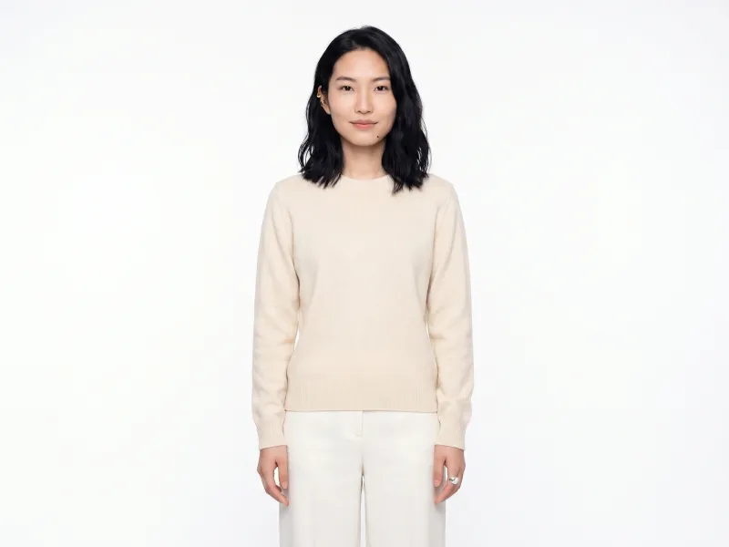 Clean e-commerce hero portrait of the same custom AI actor Mira Chen in a plain warm-cream cotton-knit crewneck against a seamless pure white infinity backdrop with even overhead key light, Playcut E-com Hero filter