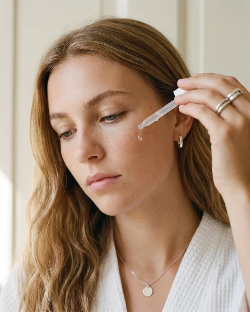 Editorial beauty still of Playcut custom AI actor Sienna Bell, a young honey-blonde model with freckles, applying a drop of clear serum to her cheekbone from a frosted-glass dropper, white waffle bathrobe, silver disc-pendant necklace, soft north-facing daylight, Playcut Actor Engine generation
