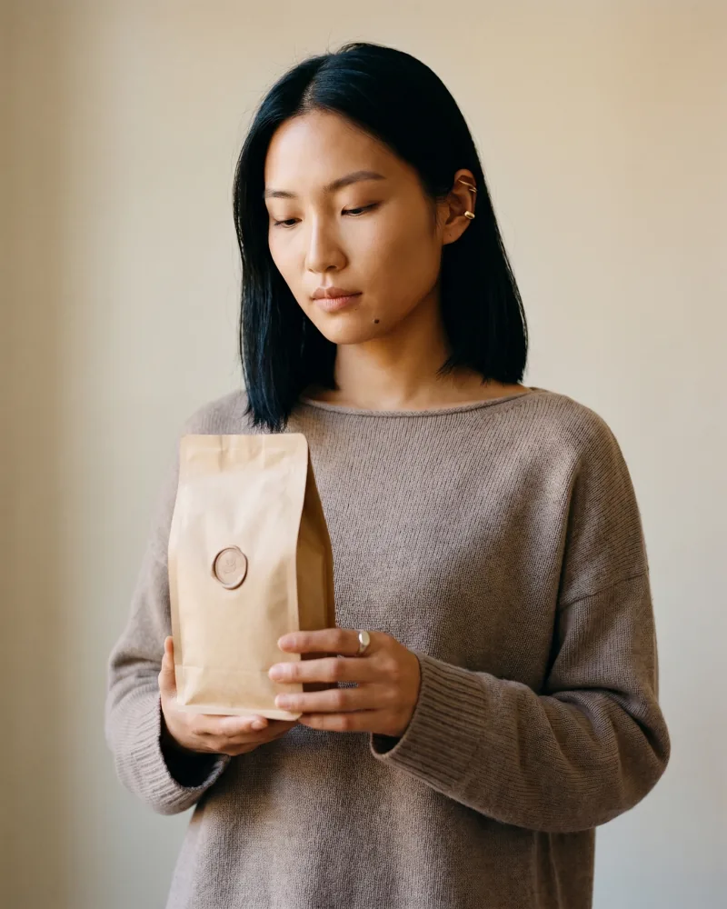 Editorial commerce still of the same custom AI actor Mira Chen holding an unbranded kraft-paper coffee bag with embossed wax-seal label, taupe boatneck merino knit, soft window light, Playcut Actor Engine generation
