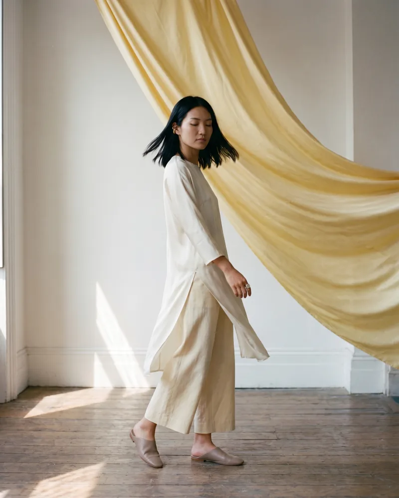 Whimsical theatrical editorial fashion photograph of the same custom AI actor Mira Chen mid-rotation in an ivory silk tunic and cream linen culottes with an enormous butter-yellow silk drape billowing diagonally behind her, painterly staging, Playcut Actor Engine generation