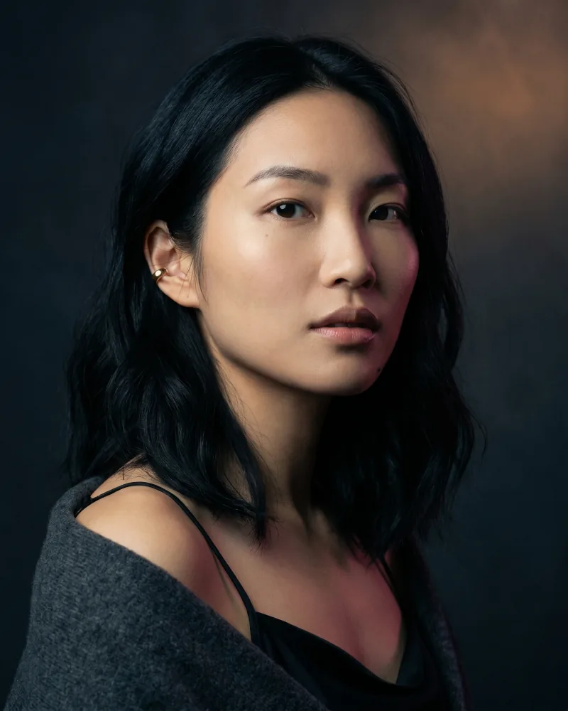 Tight close-up editorial hero portrait of the same custom AI actor Mira Chen against deep slate-charcoal velvet with theatrical Rembrandt key light, gold ear cuff visible, contemplative gaze, charcoal cashmere shawl, Playcut Actor Engine generation
