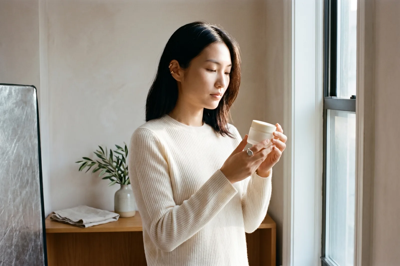 Editorial brand-shoot photograph of Playcut custom AI actor Mira Chen at a sunlit window holding an unbranded frosted-glass skincare jar at chest height, ivory cashmere ribbed crewneck, brushed-steel reflector at edge of frame suggesting an active shoot, freeze-frame of a campaign video, Playcut Actor Engine generation