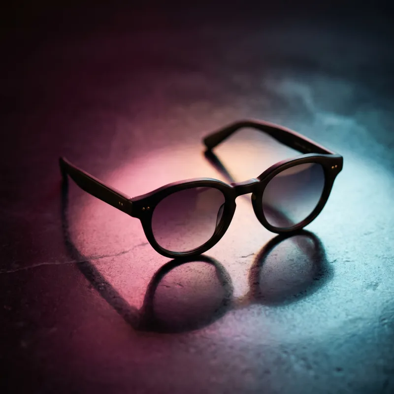 Premium nighttime editorial product still of the same matte black sunglasses on dark slate with a soft warm-magenta to cool-cyan signature gradient underglow casting a controlled colored reflection, premium nighttime aesthetic, Playcut generation