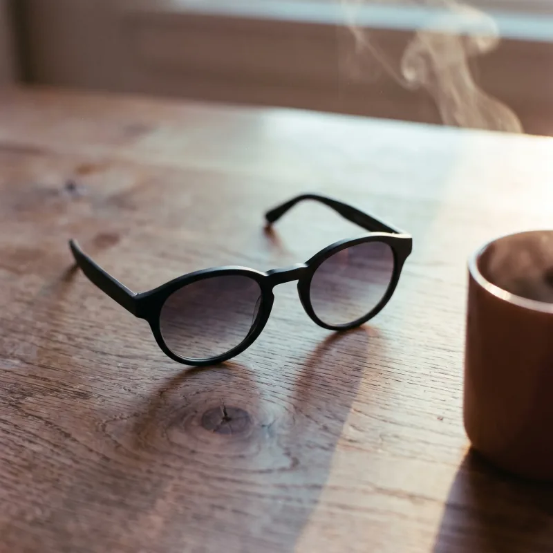 Editorial morning still-life of the same matte black sunglasses on a weathered warm-oak wood plank with a wisp of steam from a ceramic coffee mug cropped at the right edge, soft warm daylight, Playcut generation