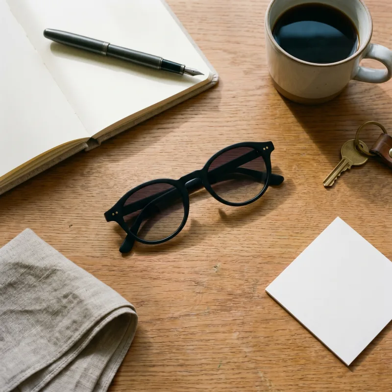 Editorial top-down flat-lay of the same matte black sunglasses on a warm-oak desk with an open linen-bound notebook, fountain pen, ceramic mug of black coffee, brass key on a leather fob and folded linen pocket square, Playcut generation