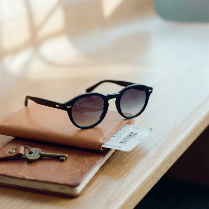 Editorial travel still-life of the same matte black sunglasses on a warm-tan natural leather passport wallet with a boarding pass peeking out, brass luggage key on a leather fob, worn travel notebook beneath, soft afternoon airport daylight, Playcut generation